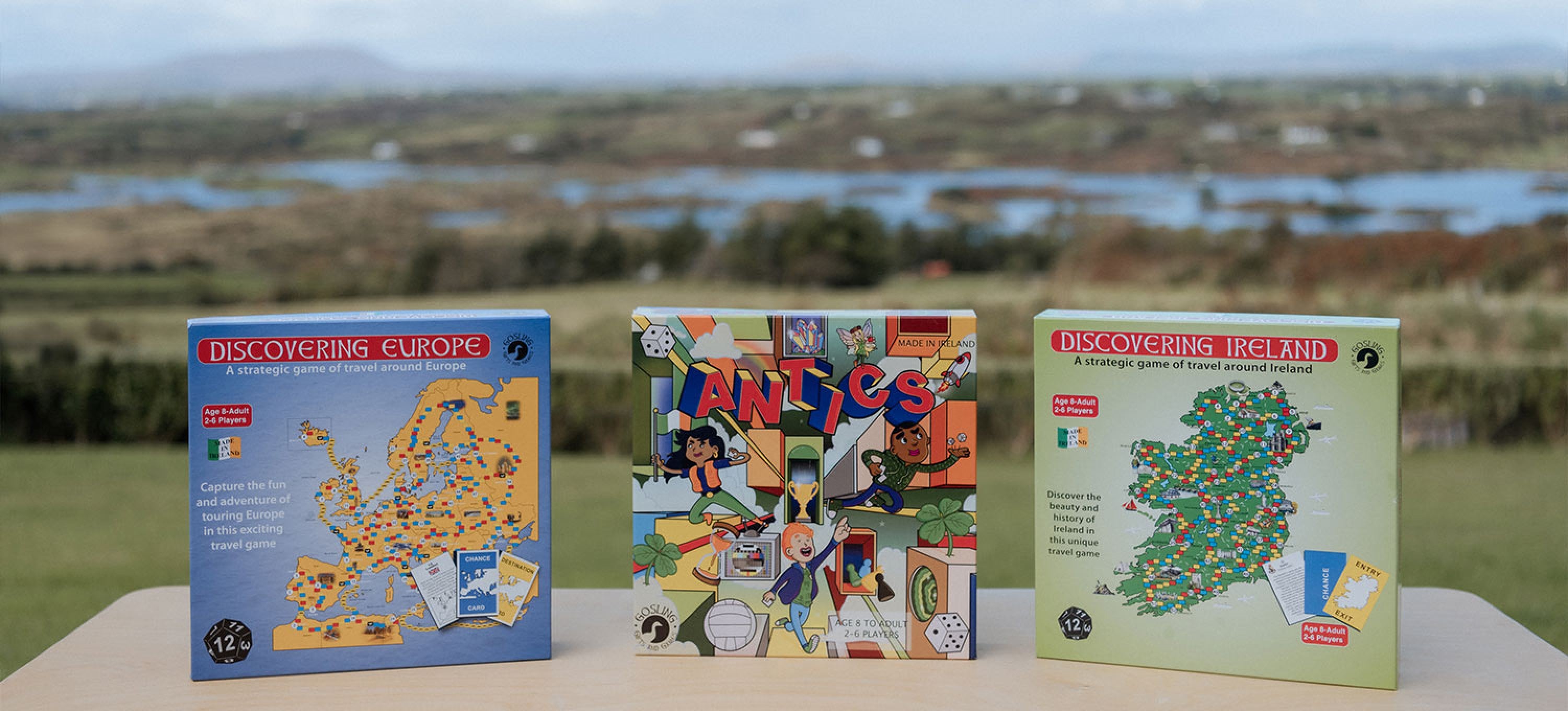 Three board game boxes standing upright on a table outdoors, with a blurred landscape of water and green fields in the background. The boxes are labeled Discovering Europe, Antiques, and Discovering Ireland.